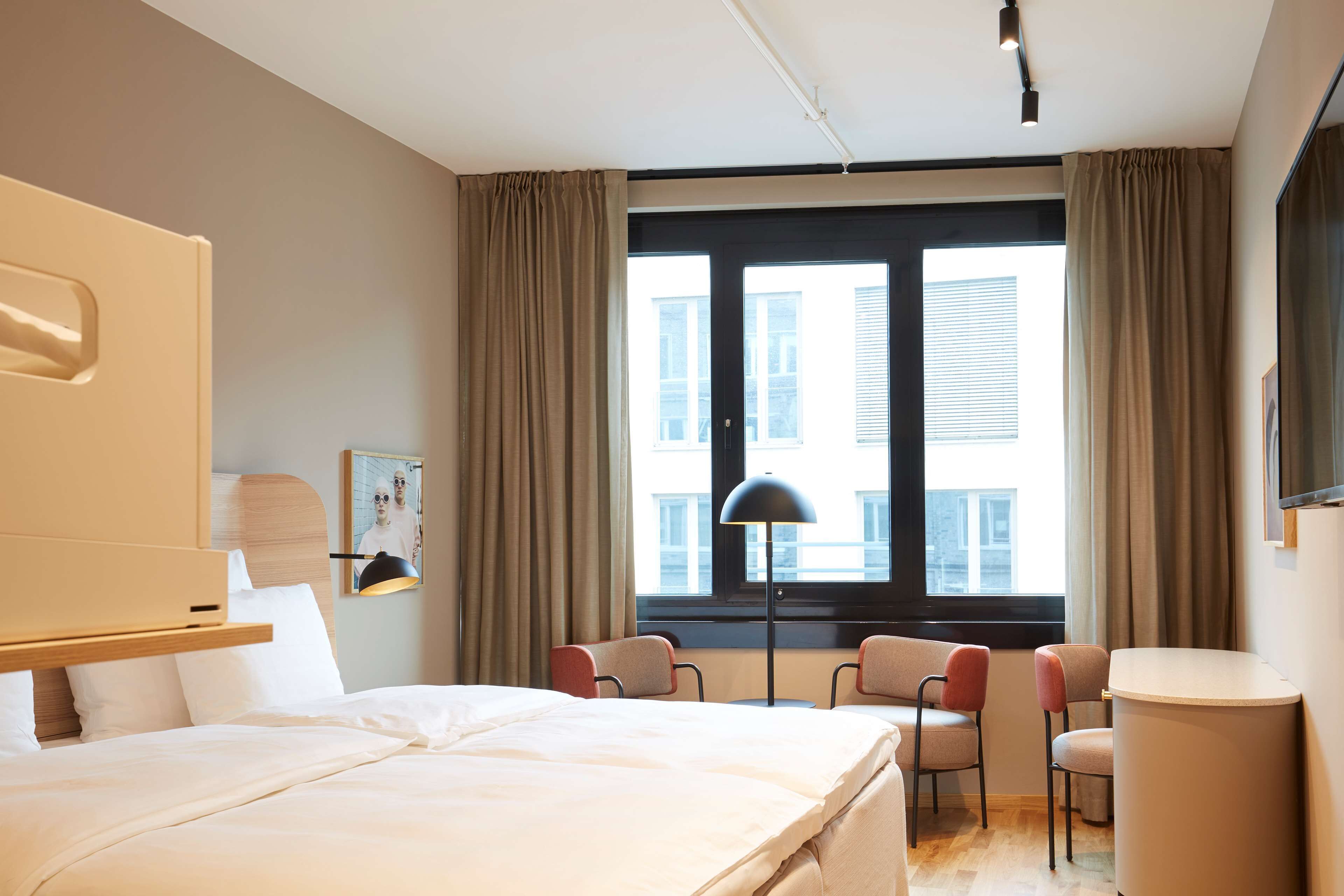 A hotel room with a bed, two chairs, a desk, a lamp, and a window with curtains.