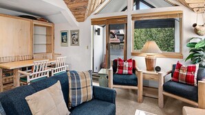 Een gezellige woonkamer met een blauwe bank, houten stoelen, een glazen salontafel en uitzicht naar buiten door een raam.