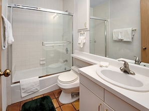 Un baño moderno con ducha de vidrio, inodoro blanco, lavamanos con espejo y alfombra verde en el suelo.