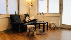 A living room with a black sofa, a small table, and two stools. There is a lamp with a flower arrangement on the wall, and a window with a view of the outside. - Alcazar by Interhome (Saas-Grund)