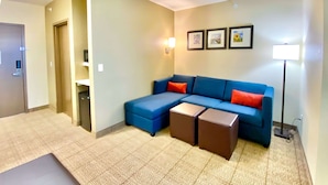 A living room with a blue sectional sofa, two brown ottomans, a patterned carpet, and a wall-mounted TV. - Comfort Inn & Suites Harrah (Harrah)