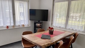 Un coin repas avec une table en bois, quatre chaises marron, un téléviseur posé sur un meuble et une fenêtre avec des rideaux.