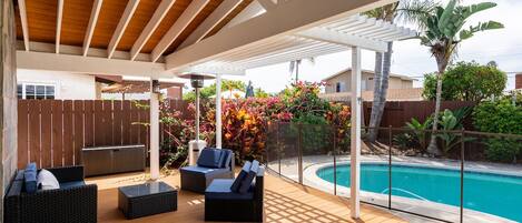 Uma área externa coberta com piso de madeira, móveis de vime preto e uma piscina.