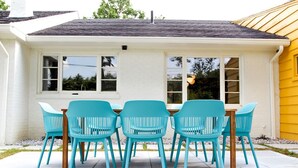 Un bâtiment blanc avec une façade jaune, un toit gris et une terrasse dotée d’une table en bois et de six chaises turquoise.