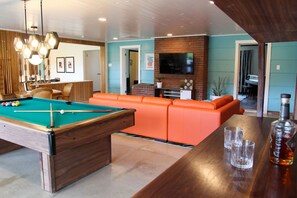 A pool table with balls, a pool cue, and a bottle of liquor on a wooden table.