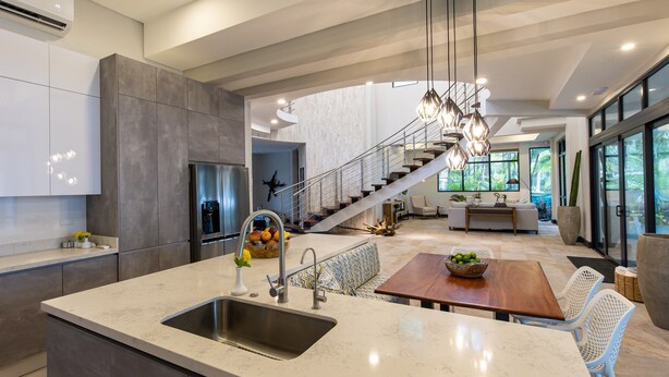 Una cocina moderna con una isla grande, electrodomésticos de acero inoxidable y una escalera que lleva a una sala de estar con sofá y mesa de comedor.