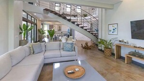 Amplia sala de estar con una escalera moderna, un sofá blanco y una mesa de centro con mandos a distancia.