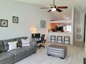 Un salon moderne avec un canapé gris, un pouf gris, un bureau avec une lampe et un ventilateur de plafond.