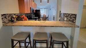 Una cocina con encimera de granito, tres taburetes de bar, armarios de madera, microondas y refrigerador.