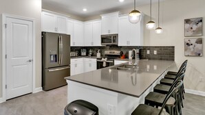 A modern kitchen with white cabinets, stainless steel appliances, a large island, and a staircase with wooden steps. - 7BD/5BA BRAND NEW Home w/pool close to Disney (Kissimmee)