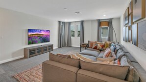 A modern living room with a large flat-screen TV, a grey sofa with multiple cushions, a small wooden coffee table, and a patterned rug. - 7BD/5BA BRAND NEW Home w/pool close to Disney (Kissimmee)