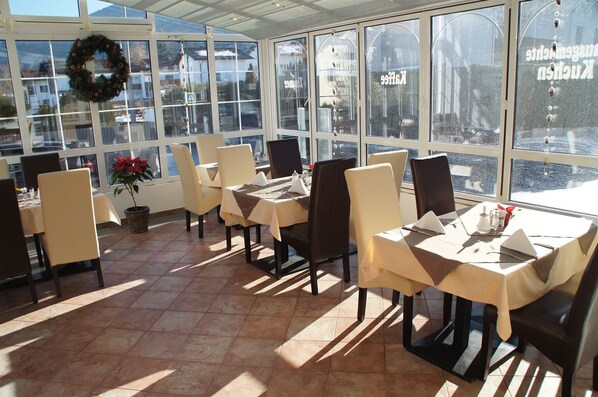 A restaurant with multiple tables and chairs, large windows, and a wreath on the wall. - Hotel Koegele (Axams)