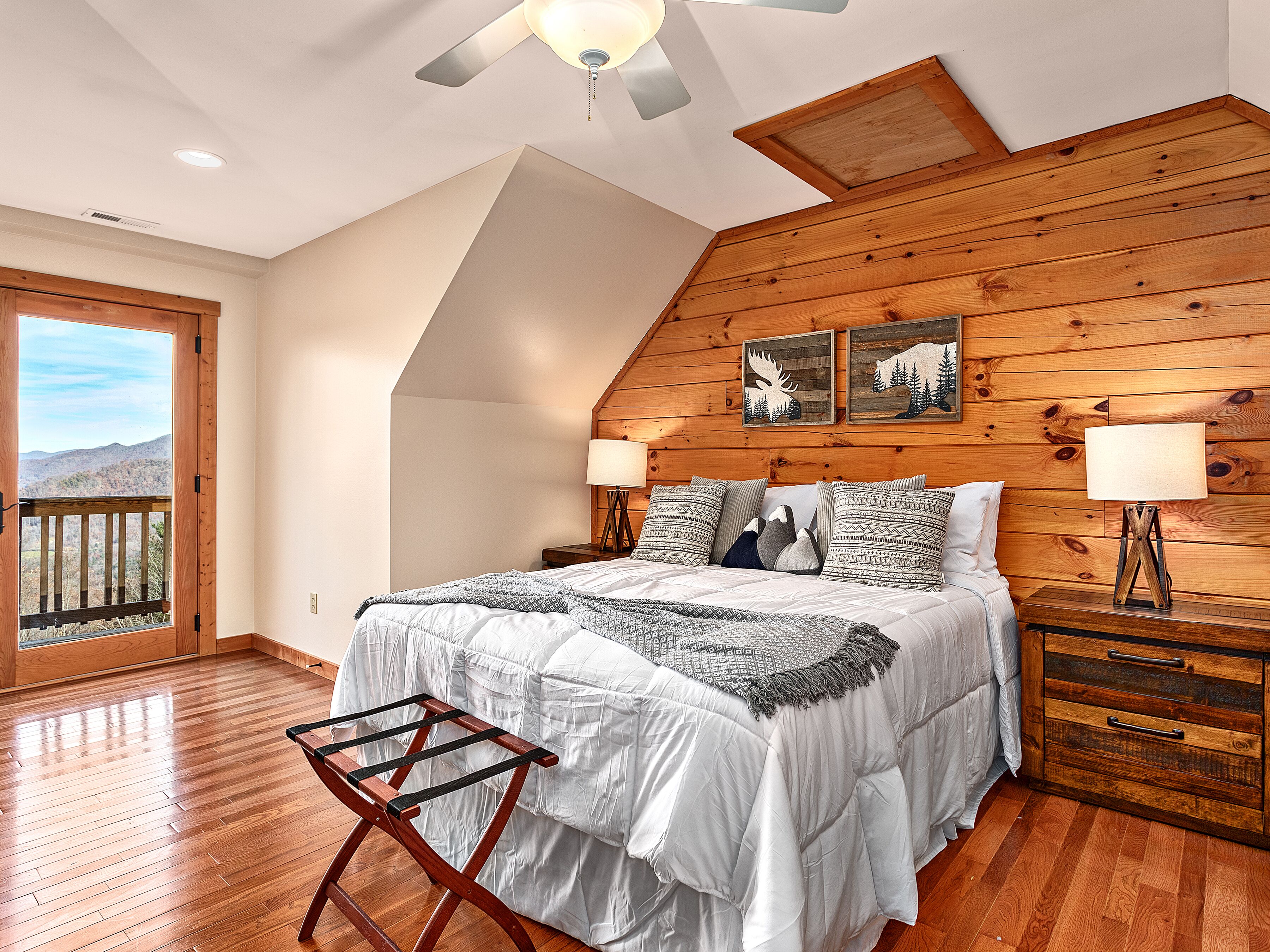 A bedroom with a sloped ceiling, wooden panel walls, a bed with white bedding, two bedside lamps, and a view through a large window.