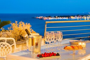A table with a champagne bottle, glasses, plates, and fruit, overlooking a coastal city at night.