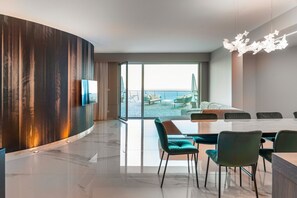 A modern hotel room with a curved wall, a large dining table, teal chairs, and a view of the beach.