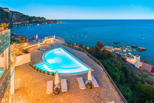 A pool with lounge chairs and umbrellas, a view of the sea, and a stone pathway.