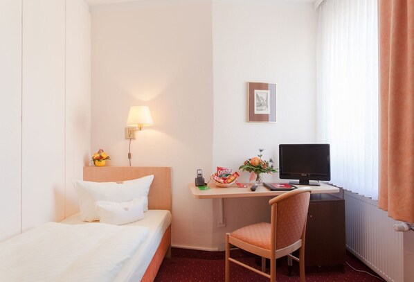 Et hotelværelse med en seng, et skrivebord med en computer, en stol, en lampe og et vindue med gardiner.
