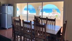 Un comedor con una mesa y sillas, un reloj en la pared y vistas al mar a través de amplios ventanales.