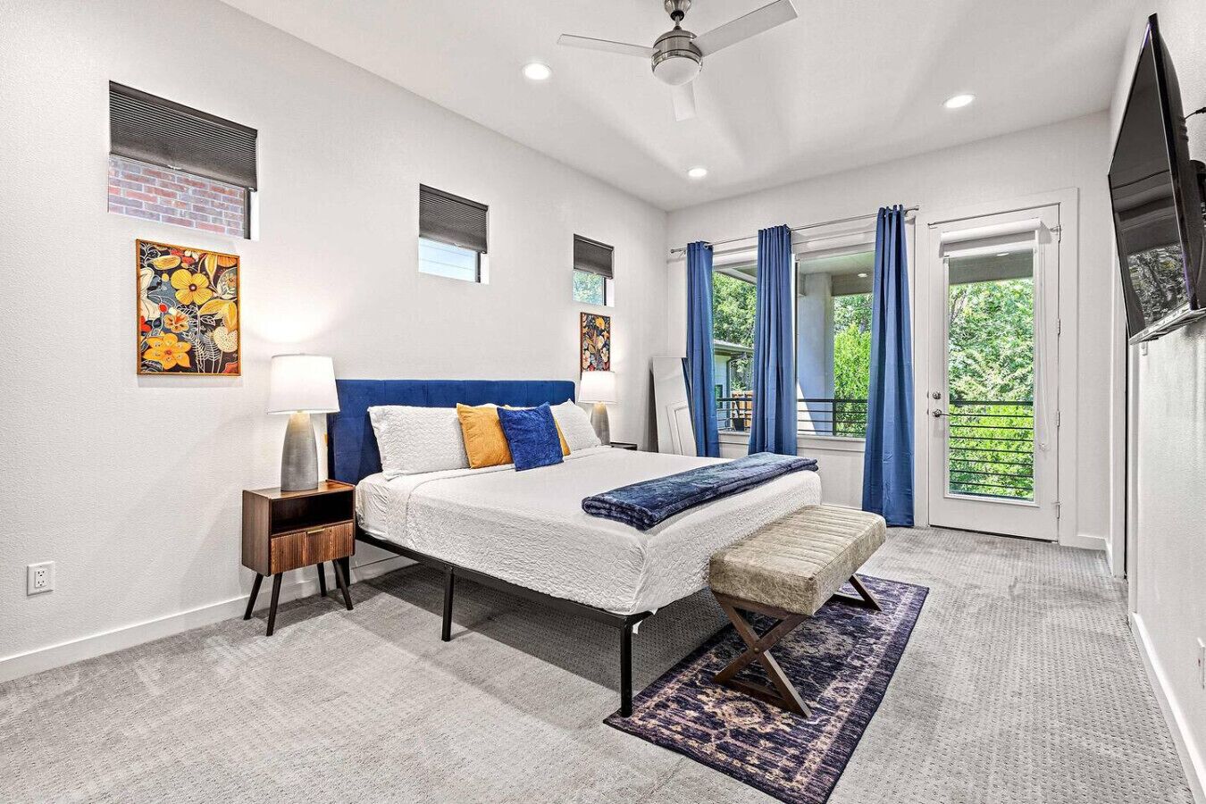 A bedroom with a bed, bedside tables, a bench, a ceiling fan, and a view of the outdoors through a sliding door.