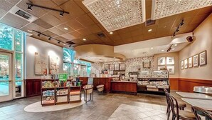 Un café espacioso con una decoración moderna, que cuenta con una barra central con una máquina de café, zona de estar con mesas y sillas, y grandes ventanales que proporcionan luz natural.