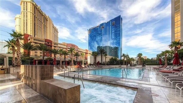 Zona de piscina de hotel con múltiples sillas de descanso y un jacuzzi, rodeada de palmeras y edificios modernos.