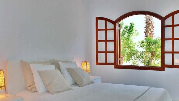 Quarto com cama, roupa de cama branca, mesa de cabeceira, luminária fixada na parede e janela com persiana aberta.