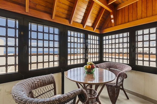 A room with a round table and wicker chairs, large windows with black frames, and a view of the beach.