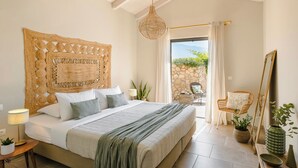 Une chambre avec un grand lit, une tête de lit en osier, un luminaire suspendu, une vue sur l’extérieur et un fauteuil avec un miroir.