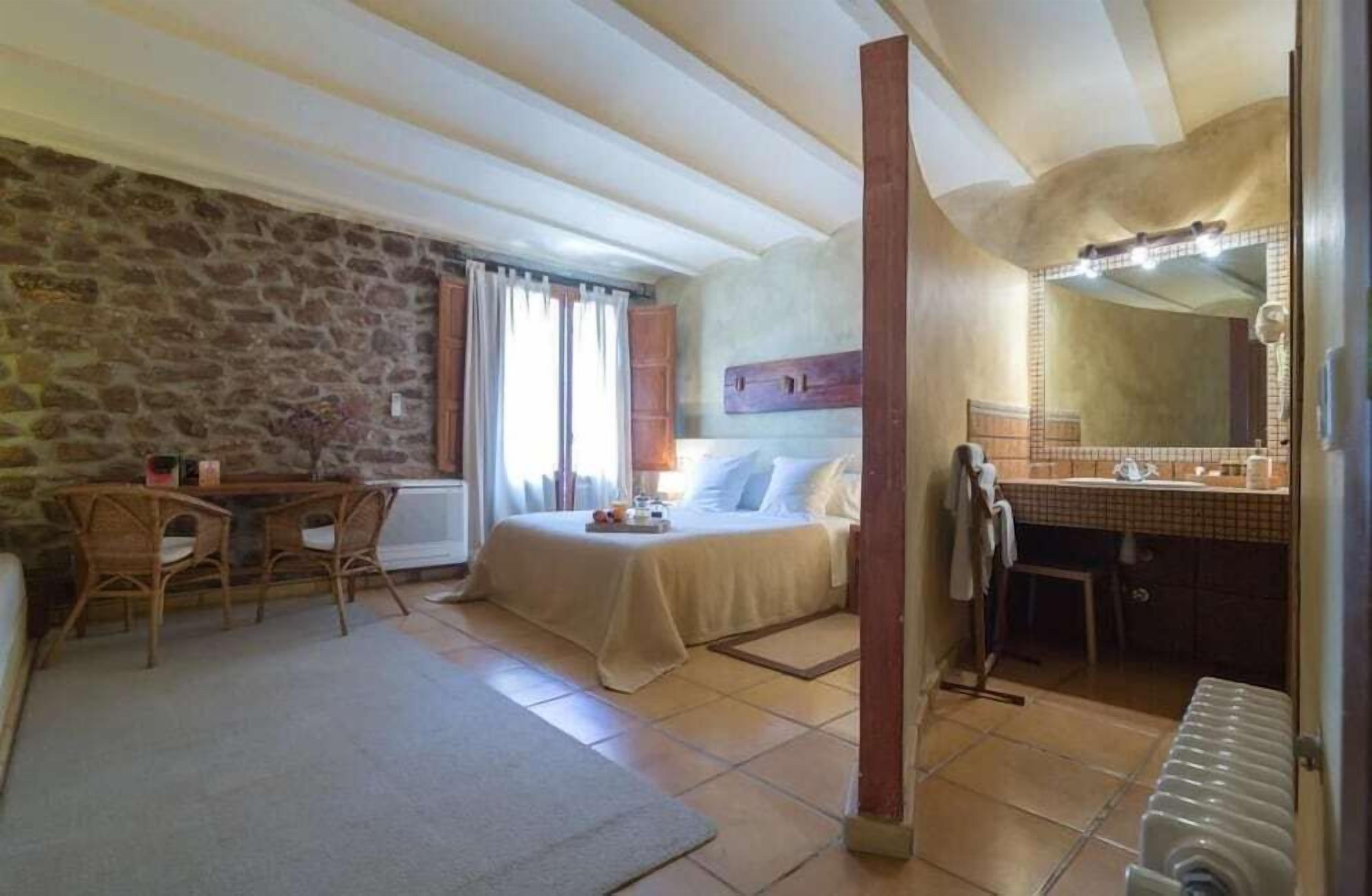 A bedroom with a stone wall, a bed, a desk, and a bathroom with a mirror and sink.