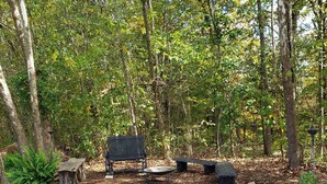 A forested area with a picnic table, a fire pit, and a stack of firewood.