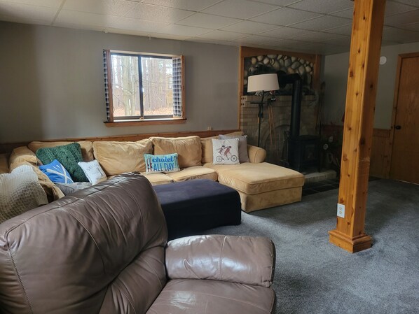 Un salon chaleureux avec un canapé beige, un pouf bleu et un divan en cuir marron. On y trouve une fenêtre avec des stores, une cheminée et un pilier en bois.