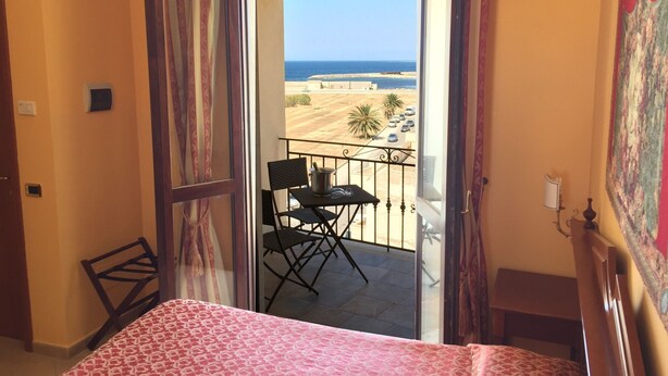 A bedroom with a balcony, a bed with a patterned bedspread, a chair, and a table with a bottle. - Hotel D Apos Angelo Palace (Mazara del Vallo)