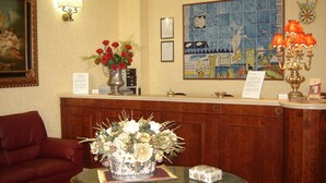 A reception area with a wooden desk, a vase of flowers, a framed painting, and a tiled wall with a cityscape. - Hotel D Apos Angelo Palace (Mazara del Vallo)
