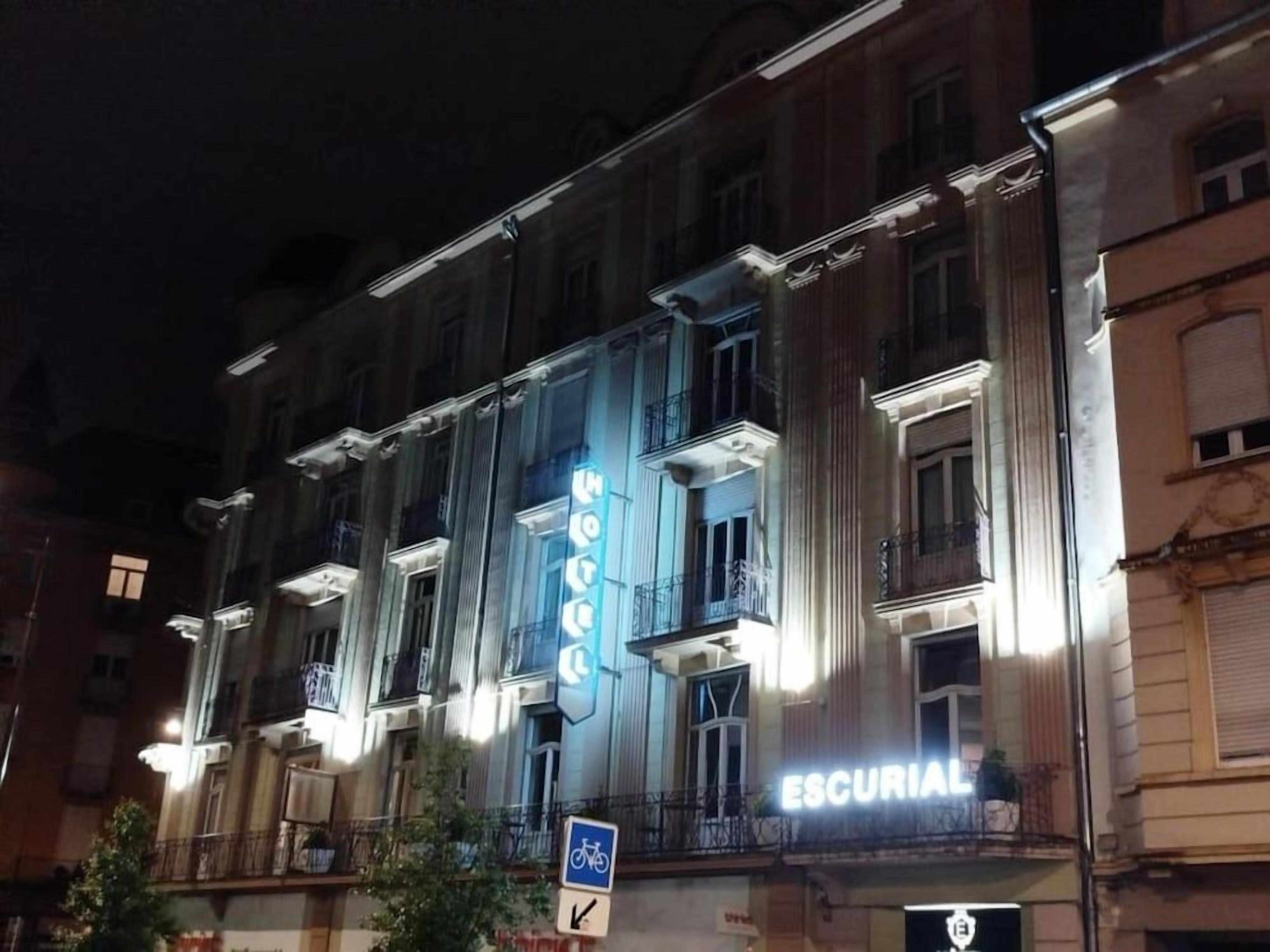 Un immeuble à plusieurs étages doté d’une enseigne lumineuse, sur laquelle on peut lire les mots « ESCURIAL » et « HÔTEL ».