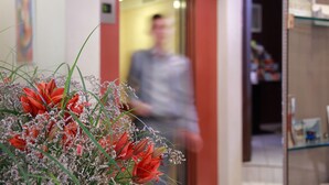 Un pasillo con una figura borrosa al fondo, una puerta de vidrio y un arreglo floral en primer plano.