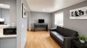 A modern living room with a sofa, a flat-screen TV on a stand, a microwave, and a small table with a plant. - Welcome Suites Hazelwood Extended Stay Hotel (Hazelwood)