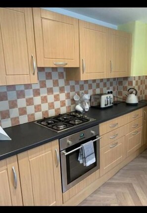 A kitchen with light wooden cabinets, a black countertop, a stove, and a tiled backsplash.