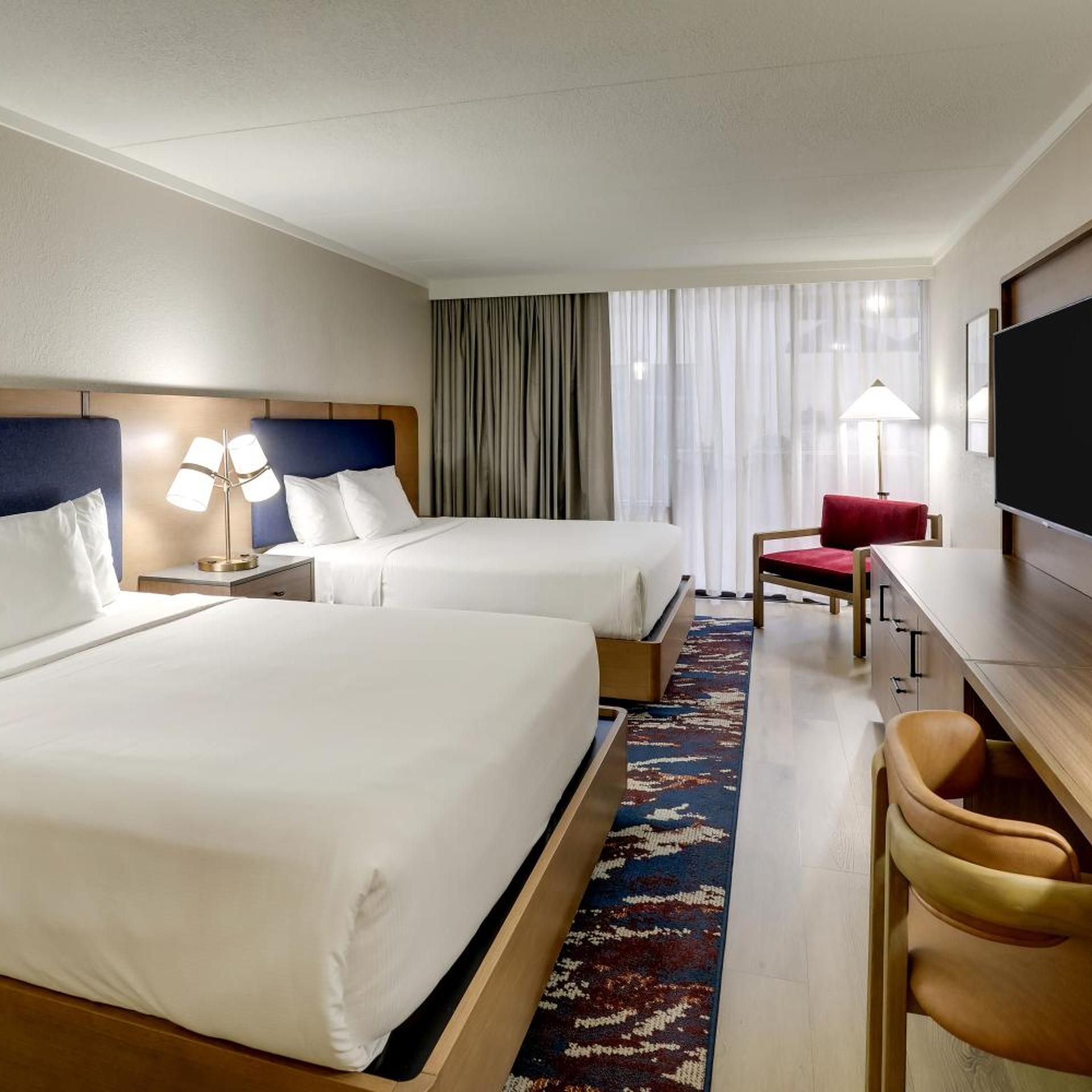 Ein Hotelzimmer mit zwei Betten, einem Schreibtisch, einem Sessel, einem Fernseher und einem Fenster mit Vorhängen.