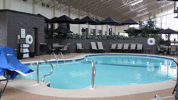 Piscina interior con sillas de descanso y sombrillas, rodeada por un techo alto y paredes con ventanas.