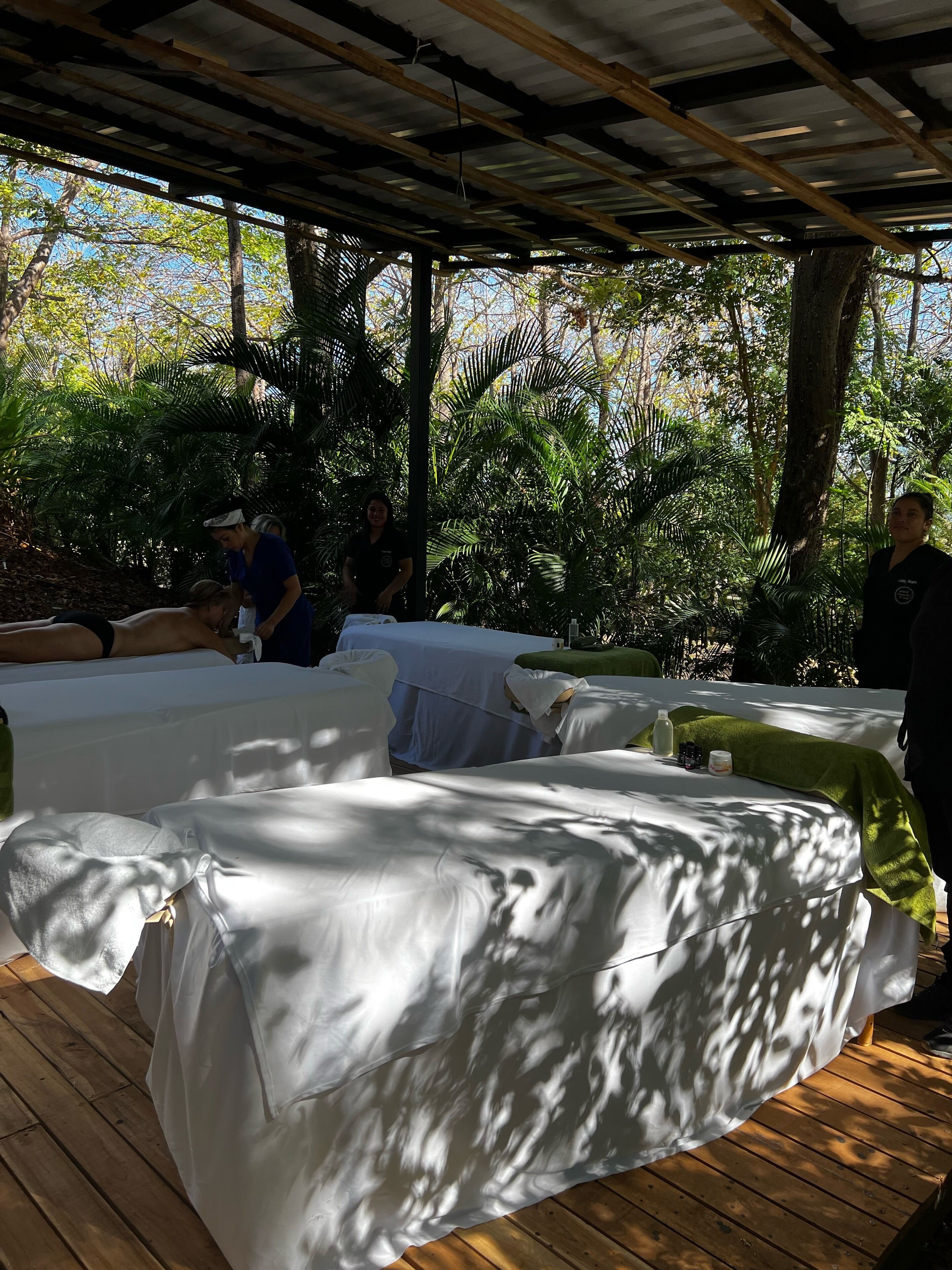 Un espace extérieur abrité, doté d’un plancher en bois et de lits blancs, entouré d’une végétation luxuriante.