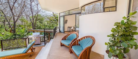 Un balcon doté d’un plancher en bois, de deux fauteuils en osier avec des coussins bleus, d’un barbecue et d’une plante en pot.