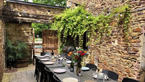 An outdoor dining area with a stone wall backdrop, a wooden deck, and a long table set for a meal. - For a vacation in the heart of the magic of Brocéliande, come and stay at Le Libert (Mauron)