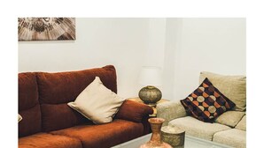 Una sala de estar con un sofá marrón, cojines beige y multicolores, una mesa de centro de vidrio con un jarrón y una lámpara sobre una mesita auxiliar.