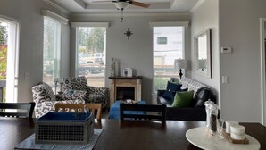 A spacious living room with a fireplace, a ceiling fan, and a large window offering a view of the outdoors. - Cooper's Cottage (Camano)