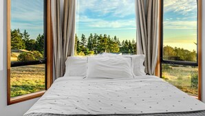 A bedroom with a large bed, two windows, and a view of a forest and fields. - Panoramic Ocean Views, Hot Tub Under The Stars and EV charging station  (Sea Ranch)