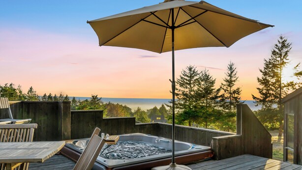 A wooden deck with a hot tub, a table, and chairs, overlooking a forested area. - Panoramic Ocean Views, Hot Tub Under The Stars and EV charging station  (Sea Ranch)