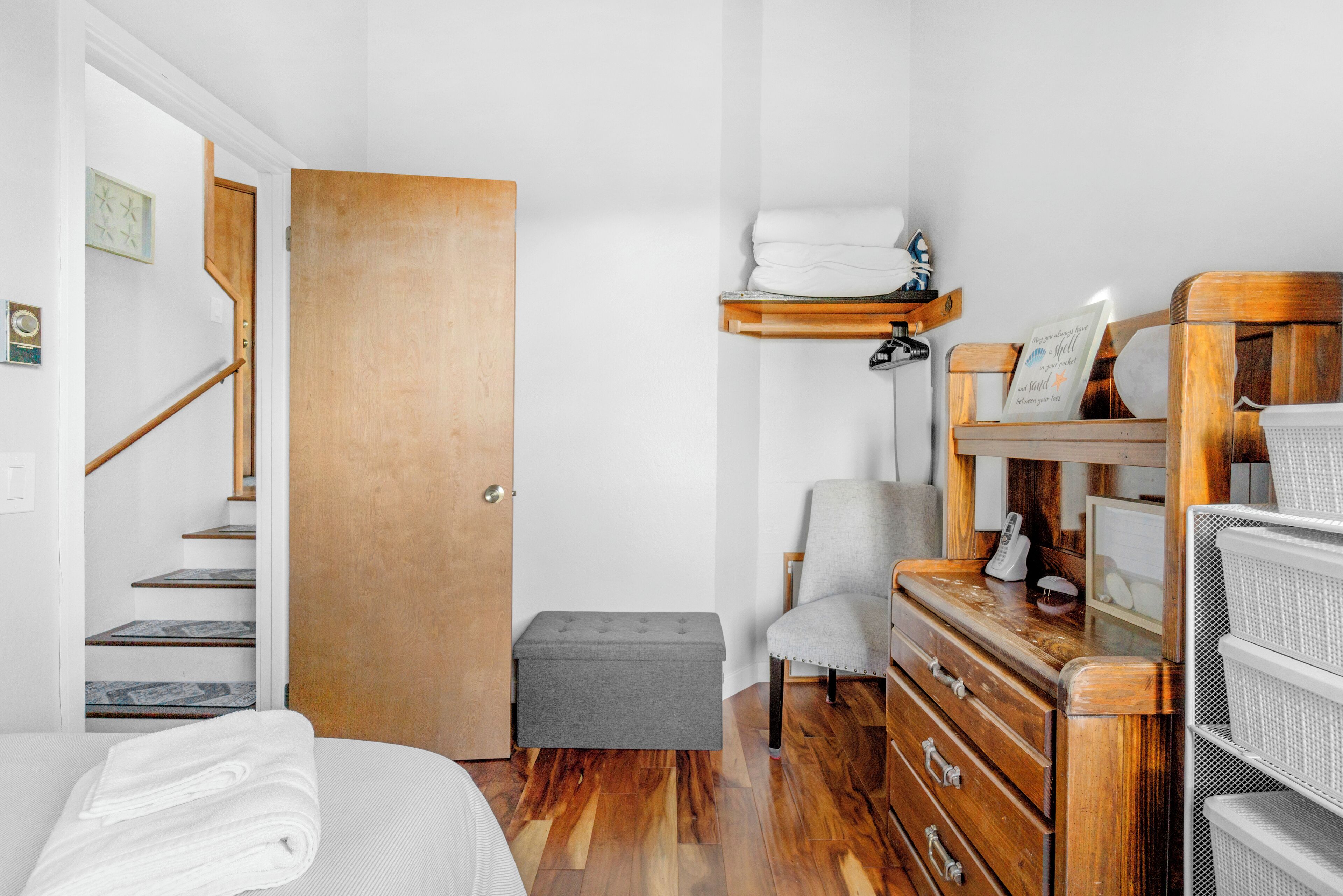 Une chambre avec une porte en bois, un banc gris, une commode en bois, une chaise et un lit avec du linge de lit blanc.