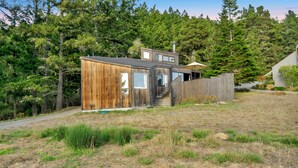A wooden cabin with a small porch and a chimney, surrounded by a grassy yard and dense forest. - Panoramic Ocean Views, Hot Tub Under The Stars and EV charging station  (Sea Ranch)