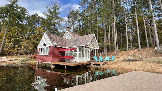 A houseboat with a covered deck and a small porch, surrounded by a forest and a body of water. - Near Munising snowmobile trails. Ride out from the property  (Munising)