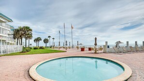 A swimming pool with a brick border, surrounded by lounge chairs and umbrellas, with palm trees and a building in the background. - Daytona Beach Condo w/ Ocean-View Balcony! (Daytona Beach)